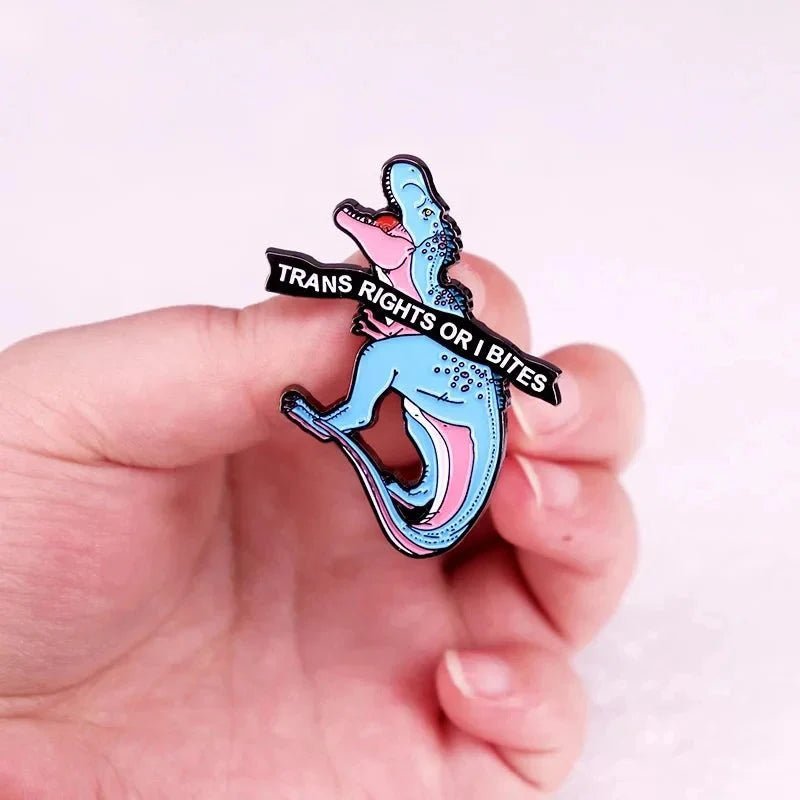 A hand holds an enamel dinosaur pin with pink accents and a black banner reading “TRANS RIGHTS OR I BITE.” The background is light and out of focus, echoing the bold message of trans rights.
