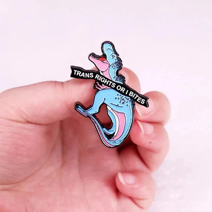 A hand holds an enamel dinosaur pin with pink accents and a black banner reading “TRANS RIGHTS OR I BITE.” The background is light and out of focus, echoing the bold message of trans rights.