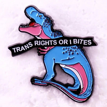 Enamel pin features a blue and pink dinosaur holding a Trans Rights or I Bites banner. Inspired by the trans pride flag, this eye-catching pin makes a bold statement for trans rights on any outfit.