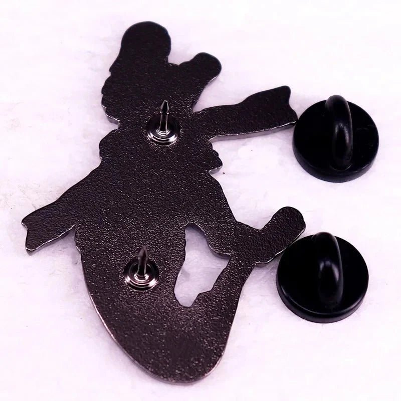 A metal pin backing shaped like a cartoon dinosaur—an enamel pin inspired by the trans pride flag—shown from behind with two pin posts and black rubber clasps, displayed on a white surface.