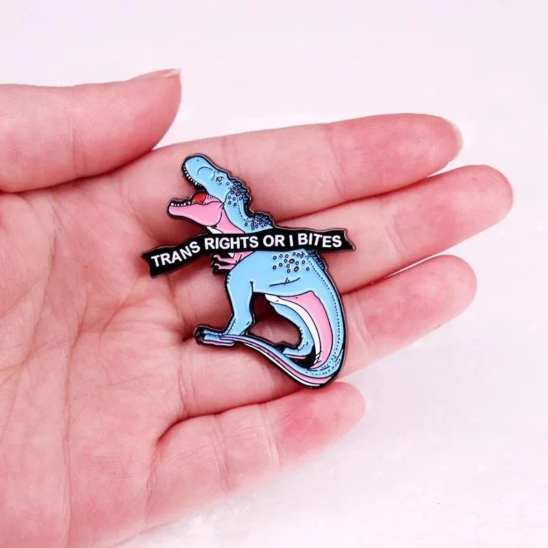 A hand holds an enamel pin, shaped like a blue and pink dinosaur with an open mouth holding a TRANS RIGHTS OR I BITES banner—an eye-catching way to show support for Trans Rights.