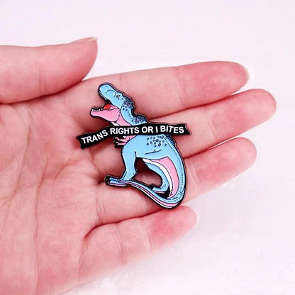 A hand holds an enamel pin, shaped like a blue and pink dinosaur with an open mouth holding a TRANS RIGHTS OR I BITES banner—an eye-catching way to show support for Trans Rights.