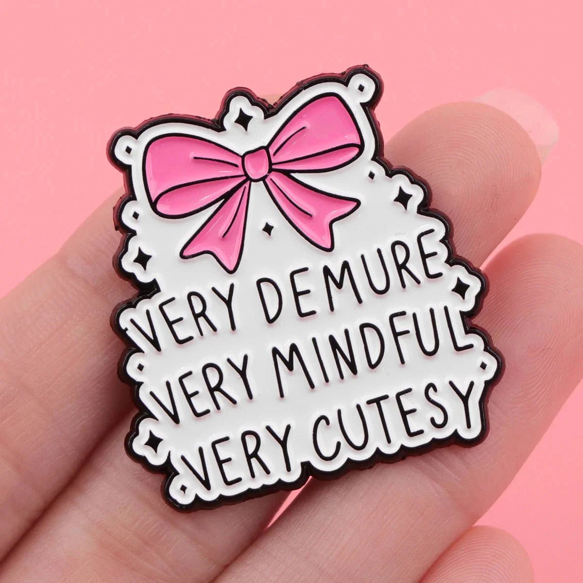 A hand holds a black and white enamel badge with a pink bow above the words Very demure, very mindful, very cutesy, on a white background with star accents, set against a pink backdrop.