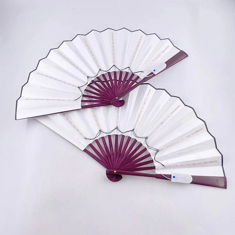 Two My Cutesy Era folding hand fans with white paper, purple edges, and purple ribbing overlap on a white background. Their elegant design is reminiscent of a battery powered LED fan, blending classic beauty with modern flair.