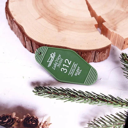 A retro green hotel key ring by Kiss Me Later, labeled Lakeview Motel 312 TOMAHAWK SHELTI HILL, rests on a white surface next to pine branches and a wooden slab.