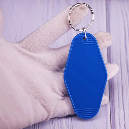 A gloved hand holds a blue, blank Kiss Me Later retro hotel key ring with a silver keyring over a light purple wooden surface.