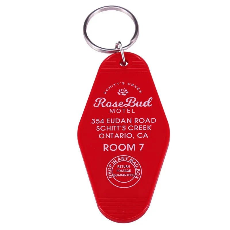A red retro hotel key ring by Kiss Me Later reads “Rosebud Motel, 354 Eudan Road, Schitt’s Creek, Ontario, CA, Room 7,” features a white rose logo and “Drop in any mailbox, return postage guaranteed.” A must-have pop culture collectible.