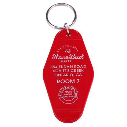 A red retro hotel key ring by Kiss Me Later reads “Rosebud Motel, 354 Eudan Road, Schitt’s Creek, Ontario, CA, Room 7,” features a white rose logo and “Drop in any mailbox, return postage guaranteed.” A must-have pop culture collectible.