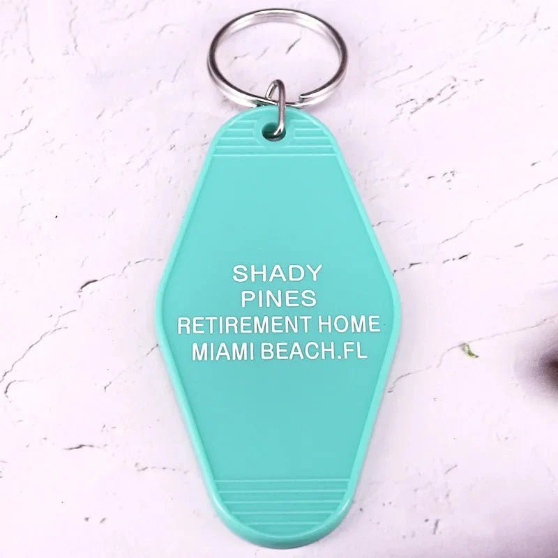 A teal, retro hotel key ring by Kiss Me Later features white text: SHADY PINES RETIREMENT HOME MIAMI BEACH, FL, set against a light, textured background.