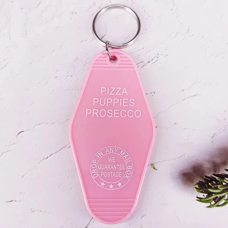 The Kiss Me Later pink retro hotel key ring features PIZZA PUPPIES PROSECCO and a stamp reading DROP IN ANY MAILBOX WE GUARANTEE POSTAGE, adding playful pop culture flair to any set of keys.