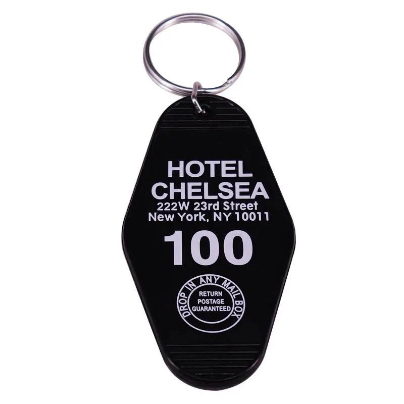 A black retro hotel key ring from Kiss Me Later with silver ring, labeled HOTEL CHELSEA, 222 W 23rd St, NY 10011 in white text and a circular emblem—RETURN POSTAGE GUARANTEED. A true pop culture collectible.