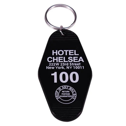 A black retro hotel key ring from Kiss Me Later with silver ring, labeled HOTEL CHELSEA, 222 W 23rd St, NY 10011 in white text and a circular emblem—RETURN POSTAGE GUARANTEED. A true pop culture collectible.