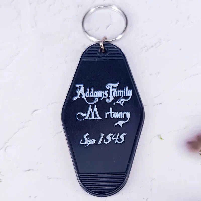 A black coffin-shaped keychain by Kiss Me Later, inspired by retro hotel key rings, features white Addams Family Mortuary, Since 1543 text and is displayed on a light-colored surface.