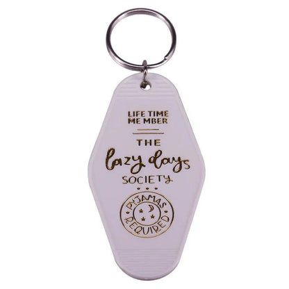 A white retro hotel key ring by Kiss Me Later features gold text: LIFE TIME MEMBER THE lazy days SOCIETY PAJAMAS REQUIRED, with a pajamas and crescent moon design—ideal for pop culture fans and lovers of unique, colorful key rings.