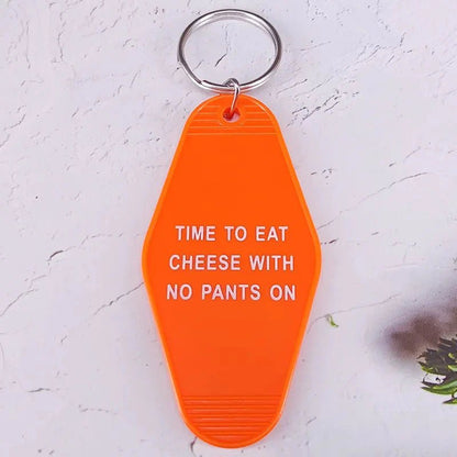 A vibrant orange retro hotel key ring by Kiss Me Later, featuring bold white text TIME TO EAT CHEESE WITH NO PANTS ON, rests on a textured surface—perfect for adding playful pop culture flair to your keys.