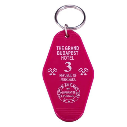 A pink retro hotel key ring by Kiss Me Later with white text reading The Grand Budapest Hotel, 3, Republic of Zubrowka—featuring key images and a guarantee stamp. Perfect for pop culture fans and lovers of colourful key rings.
