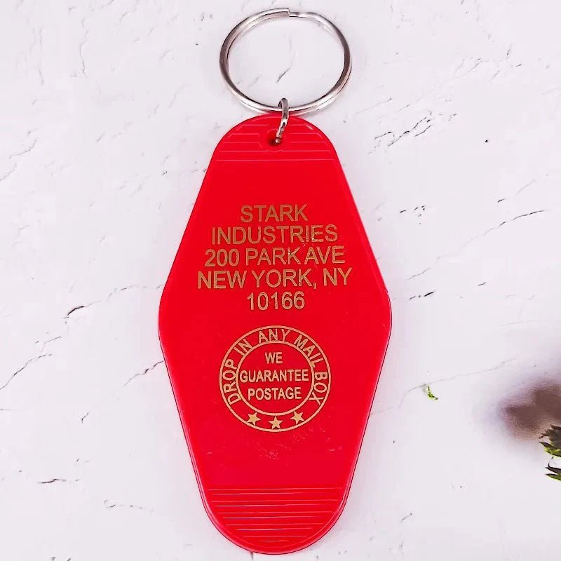 A red Kiss Me Later pop culture keychain tag with gold STARK INDUSTRIES, 200 PARK AVE, NEW YORK, NY 10166 text and a circular stamp: DROP IN ANY MAIL BOX, WE GUARANTEE POSTAGE.