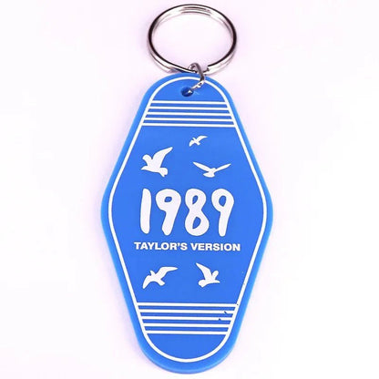 A blue retro hotel key ring by Kiss Me Later, featuring white birds, stripes, and 1989 TAYLORS VERSION in bold white letters—a stylish pop culture accessory.