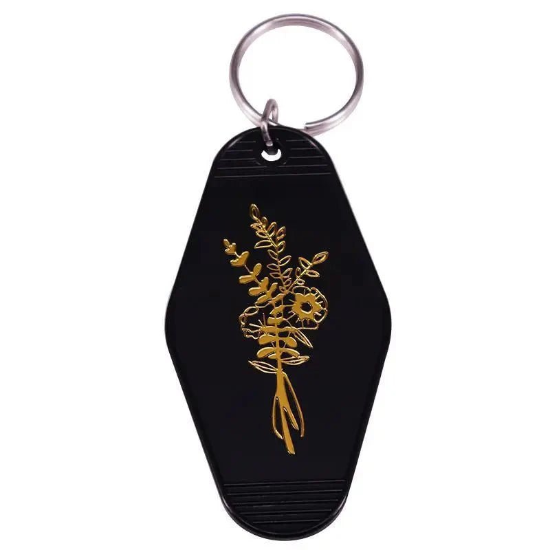 A black hexagonal keychain from Kiss Me Later with a metal ring and a gold floral design—a stylish nod to vintage hotel key rings.