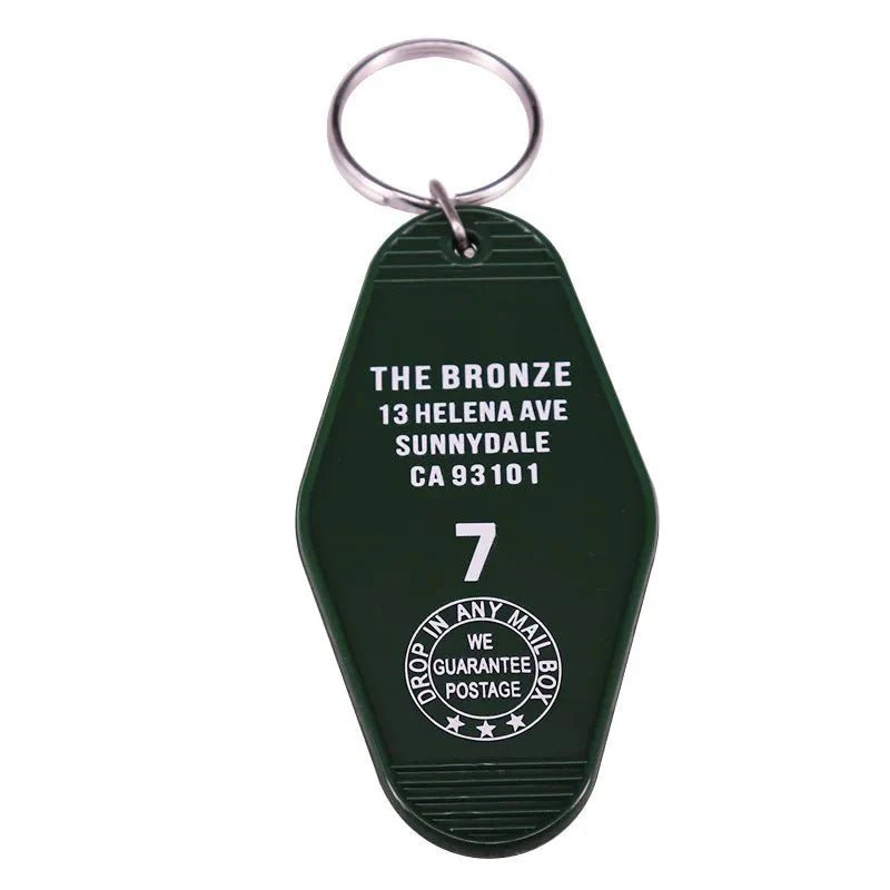 A dark green retro hotel key ring by Kiss Me Later, featuring white text: THE BRONZE, 13 HELENA AVE, SUNNYDALE, CA 93101, 7 and a round emblem with DROP IN ANY MAIL BOX, WE GUARANTEE POSTAGE—a classic pop culture nod.