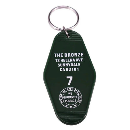 A dark green retro hotel key ring by Kiss Me Later, featuring white text: THE BRONZE, 13 HELENA AVE, SUNNYDALE, CA 93101, 7 and a round emblem with DROP IN ANY MAIL BOX, WE GUARANTEE POSTAGE—a classic pop culture nod.