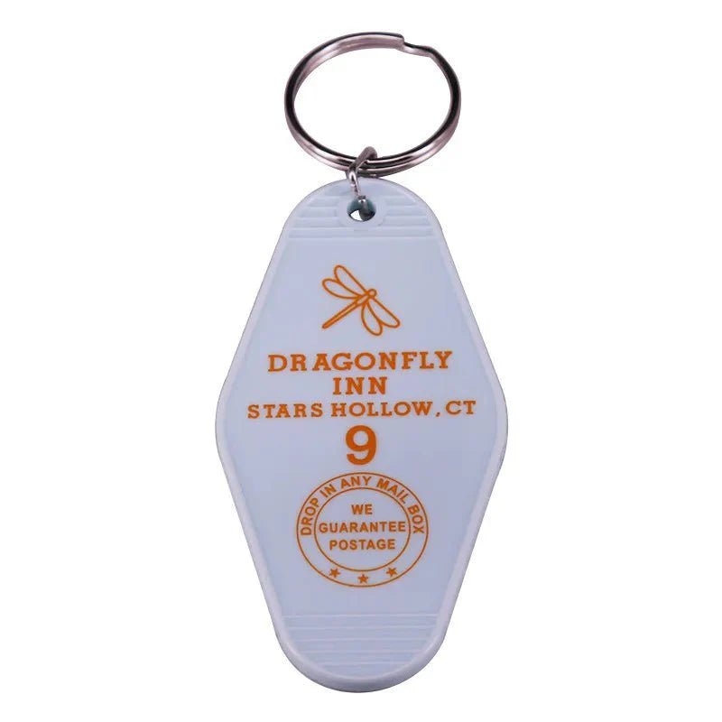 Inspired by retro hotel key rings, this white motel-style keychain from Kiss Me Later features a colorful dragonfly graphic, DRAGONFLY INN, STARS HOLLOW, CT text, and a unique DROP IN ANY MAIL BOX circular stamp for pop culture fans.