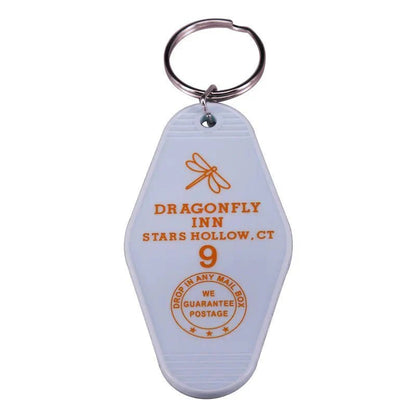 Inspired by retro hotel key rings, this white motel-style keychain from Kiss Me Later features a colorful dragonfly graphic, DRAGONFLY INN, STARS HOLLOW, CT text, and a unique DROP IN ANY MAIL BOX circular stamp for pop culture fans.