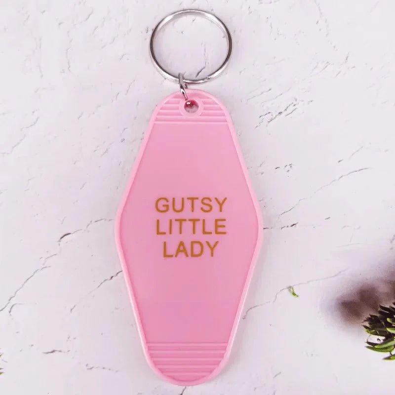 A pink, retro hotel key ring from Kiss Me Later features gold text reading GUTSY LITTLE LADY, displayed on a light background with subtle plant details.