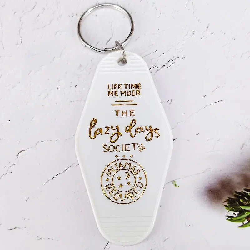 A white retro hotel key ring by Kiss Me Later with gold embossed text: LIFE TIME MEMBER THE lazy days SOCIETY PYJAMAS REQUIRED, featuring a small moon and stars illustration at the bottom.