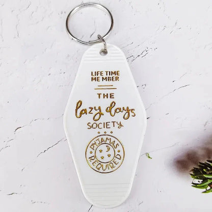 A white retro hotel key ring by Kiss Me Later with gold embossed text: LIFE TIME MEMBER THE lazy days SOCIETY PYJAMAS REQUIRED, featuring a small moon and stars illustration at the bottom.