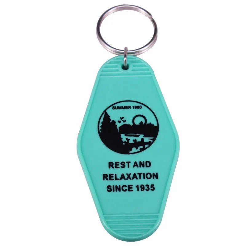 The Kiss Me Later teal retro hotel key ring has a vintage motel shape with trees, a bridge, and Ferris wheel graphic. It features the text SUMMER 1980 and REST AND RELAXATION SINCE 1935, evoking pop culture nostalgia.