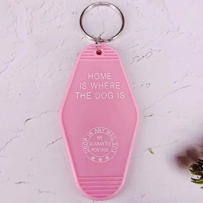 A pink retro hotel key ring by Kiss Me Later with white text: HOME IS WHERE THE DOG IS, plus a circular stamp below that says DROP IN ANY MAIL BOX - WE GUARANTEE POSTAGE.