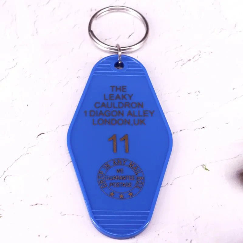 A blue vintage-style key tag by Kiss Me Later is inspired by pop culture and reads: THE LEAKY CAULDRON, 1 DIAGON ALLEY, LONDON, UK, 11. It has a mailing stamp design and comes with a metal key ring.