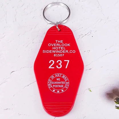 A red retro hotel key ring by Kiss Me Later features white text: The Overlook Hotel, Sidewinder, CO 81507, 237, plus a circular Drop in any mailbox, we guarantee postage stamp—perfect for pop culture fans and collectors.