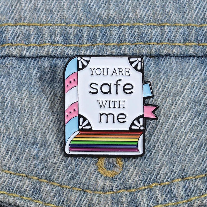 A Kiss Me Later enamel pin shaped like a book, with You are safe with me text and trans & rainbow pride colors, is attached to blue denim fabric.