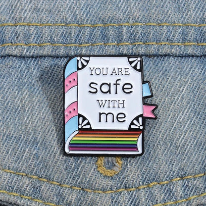 A Kiss Me Later enamel pin shaped like a book, with You are safe with me text and trans & rainbow pride colors, is attached to blue denim fabric.