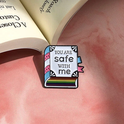 A Kiss Me Later enamel pin, shaped like a book and reading YOU ARE SAFE WITH ME, sits on a soft pink surface beside an open book, proudly celebrating trans pride.