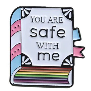 A pastel enamel pin by Kiss Me Later shaped like a book with blue, pink, and rainbow stripes for pride. The cover says YOU ARE safe WITH me, and pastel blue and pink ribbon bookmarks in trans pride colors peek out from the side.