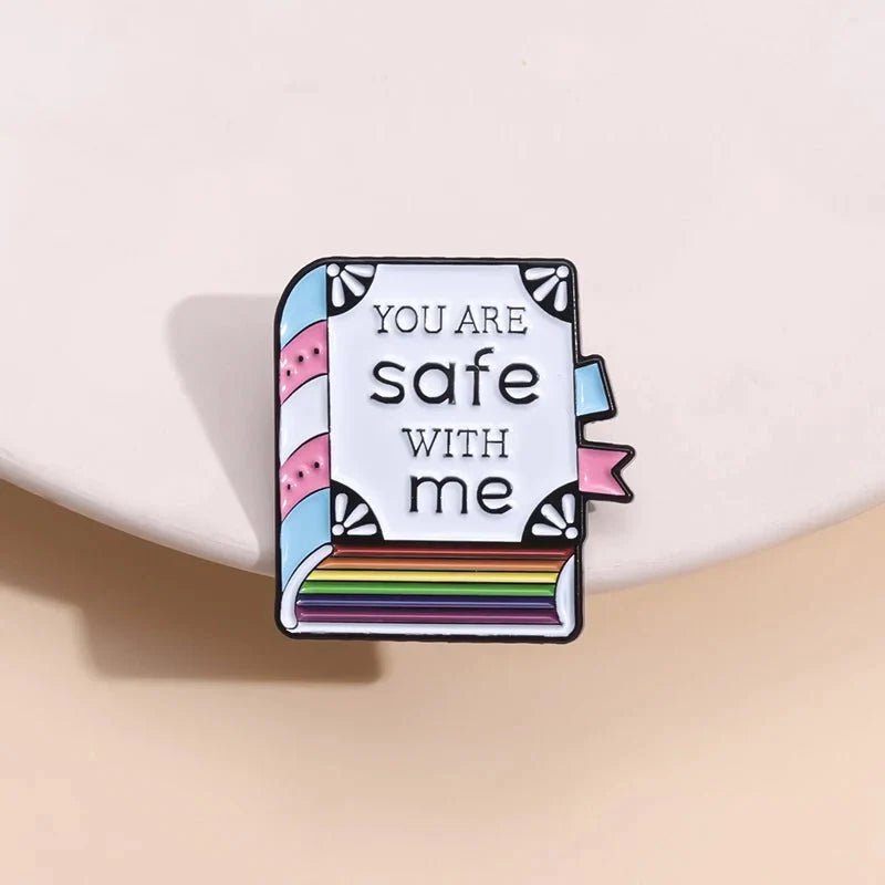 Kiss Me Later’s enamel pin features a book with rainbow-striped pages, “You are safe with me” text, and trans pride colors—blue, pink, and white—set against a light beige background.