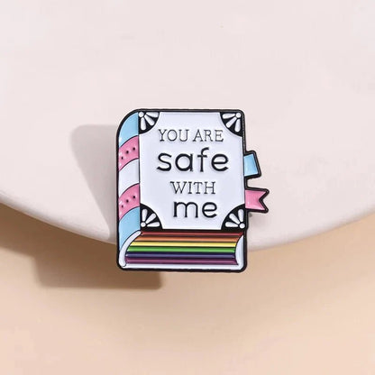 Kiss Me Later’s enamel pin features a book with rainbow-striped pages, “You are safe with me” text, and trans pride colors—blue, pink, and white—set against a light beige background.