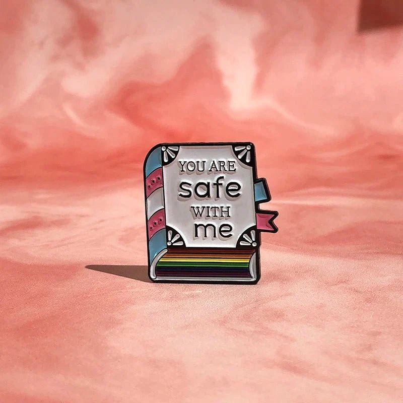 A colorful enamel pin by Kiss Me Later, shaped like a book, rests on pink fabric. Inspired by trans pride, the pin reads YOU ARE safe WITH me in bold with heart and ribbon details on the spine and pages.