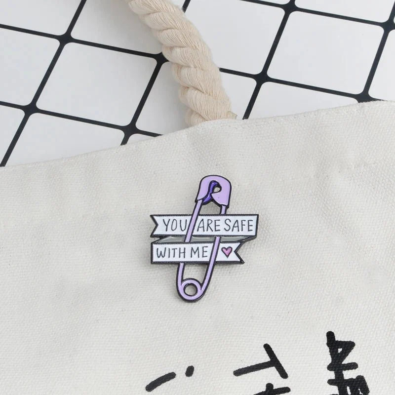 A purple safety pin enamel pin by Gay Skate is attached to a cream-colored fabric bag. Featuring two banners with YOU ARE SAFE WITH ME and a small pink heart, its a perfect pride accessory for showing support and care.