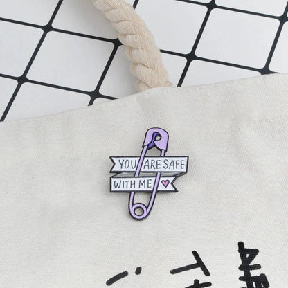 A purple safety pin heart enamel badge by Gay Skate, with banners reading YOU ARE SAFE WITH ME and a pink heart, is pinned to a white fabric bag on a tiled background.