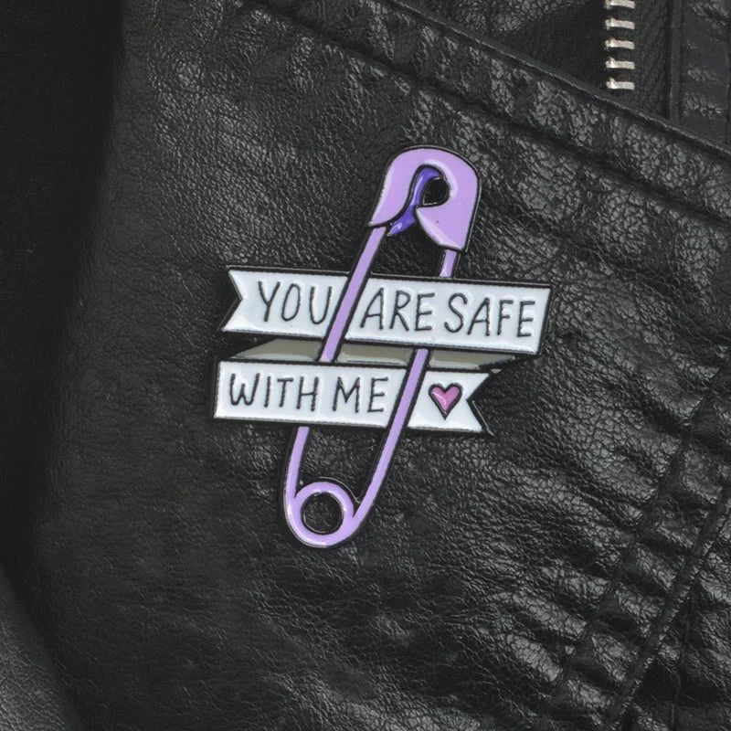 A purple safety pin enamel pin by Gay Skate on a black leather jacket reads YOU ARE SAFE WITH ME, paired with a small pink heart badge—ideal for those seeking meaningful pride accessories.