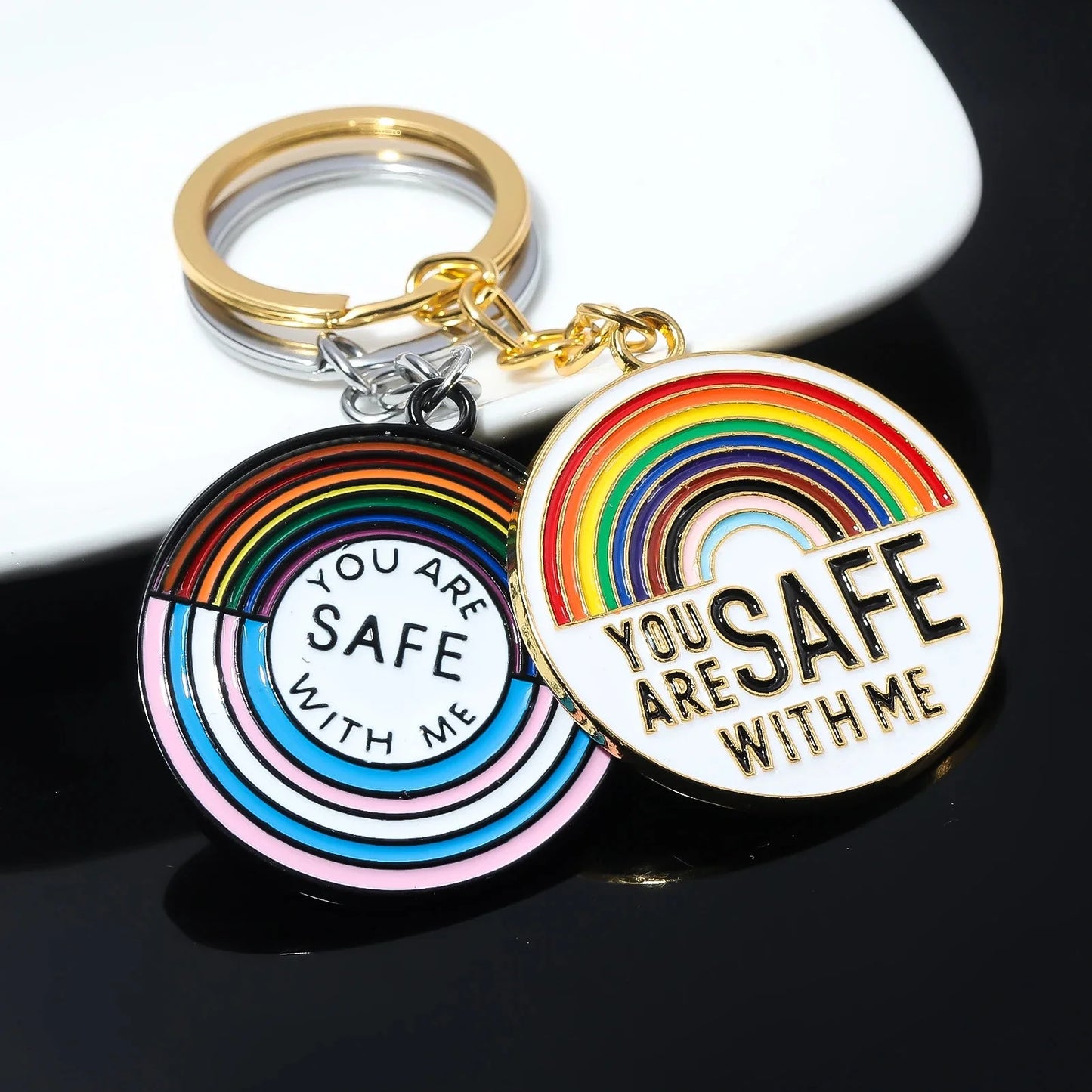 Two round keychains feature rainbow designs with YOU ARE SAFE WITH ME—one in pastel, one in bold colors. Both come with gold-colored key rings. Enjoy worldwide shipping on these heartfelt accessories!.
