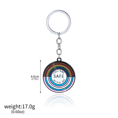 A round rainbow-striped keychain  with You Are Safe With Me in the center, attached to a silver keyring. Inspired by trans pride, it measures 4 cm and weighs 17 g. Worldwide shipping available.