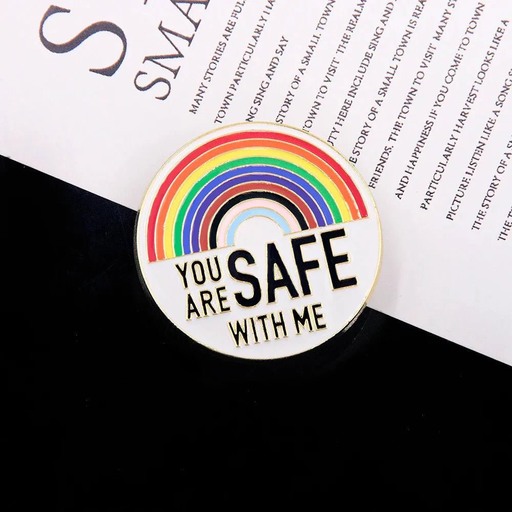 A circular pin with a rainbow design and YOU ARE SAFE WITH ME is shown on a black and white background, next to other rainbow pride enamel badges. Ships worldwide.