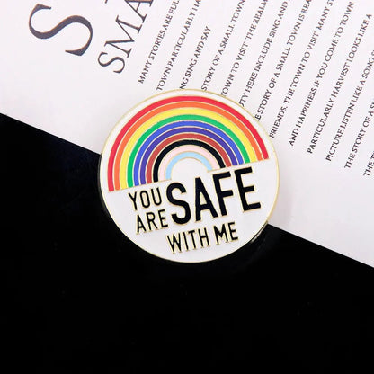 A circular pin with a rainbow design and YOU ARE SAFE WITH ME is shown on a black and white background, next to other rainbow pride enamel badges. Ships worldwide.
