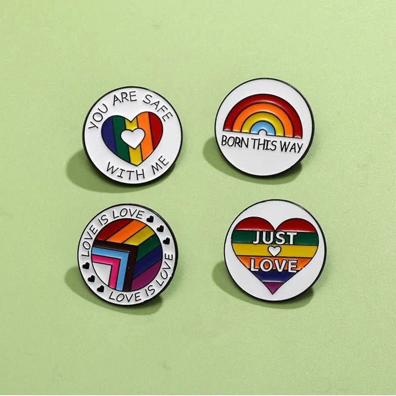 Four round rainbow pride enamel pins  on a light green background, featuring LGBTQ+ designs: rainbow hearts and messages YOU ARE SAFE WITH ME, BORN THIS WAY, LOVE IS LOVE, and JUST LOVE. Ships worldwide.