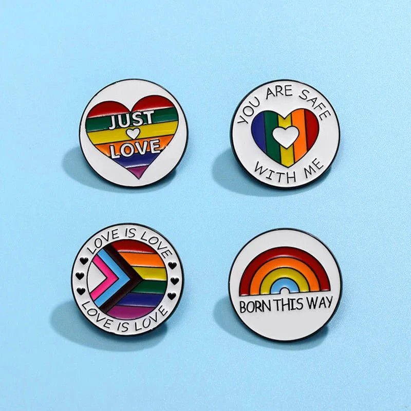Four round pins on a light blue background: rainbow heart “Just Love,” rainbow heart “You Are Safe With Me,” chevron pride heart “Love is Love,” and rainbow enamel pin “Born This Way.” Ships worldwide.